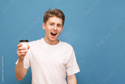 Portrait of an emotional guy with a cup of coffee in his hands, wearing a white T-shirt, looking into the camera. Copyspace