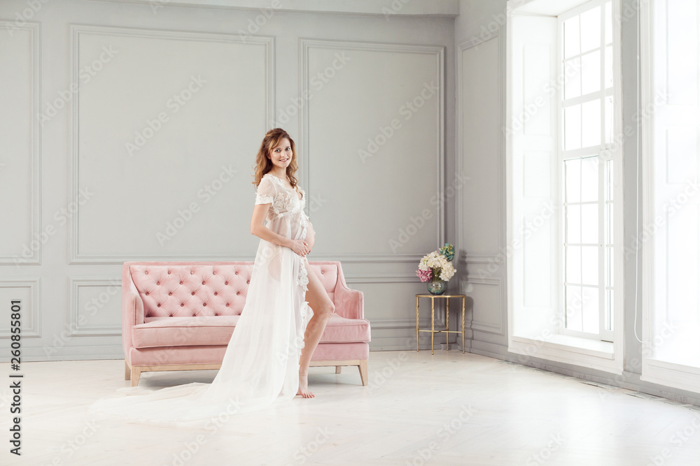 Full length portrait of beautiful pregnant young woman in white dress peignoir standing near pink sofa, holding with love her belly. Last months pregnancy, studio shot, looking at camera, toothy smile Full length portrait of beautiful pregnant young woman in white dress peignoir standing near pink sofa, holding with love her belly. Last months pregnancy, studio shot, looking at camera, toothy smile