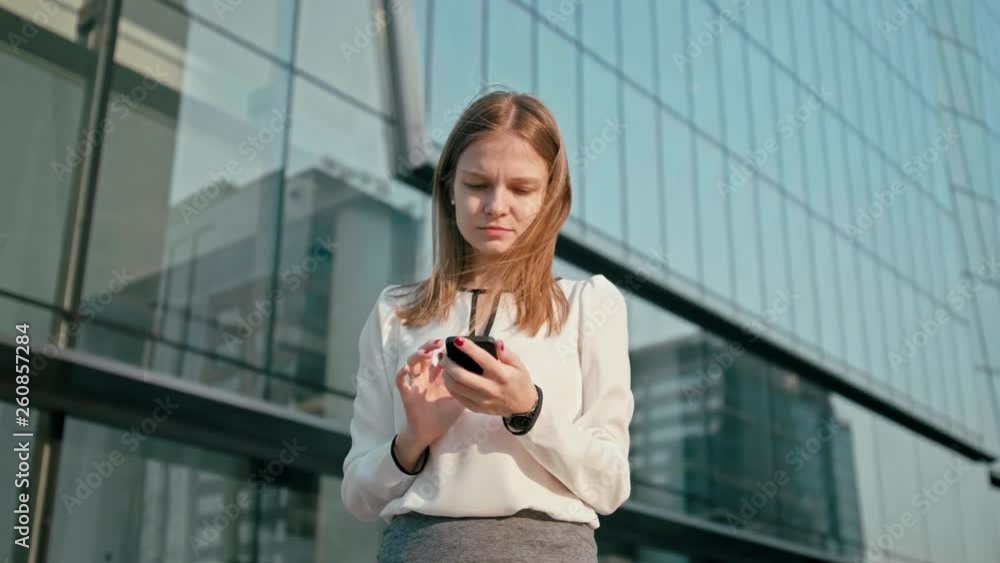 Adult Caucasian Confident Young Business Woman is Using Smartphone App Outside near Modern Office Building. Medium Low Angle 4K Slow Motion Corporate Shot with Moving Camera Around Adult Caucasian Confident Young Business Woman is Using Smartphone App Outside near Modern Office Building. Medium Low Angle 4K Slow Motion Corporate Shot with Moving Camera Around