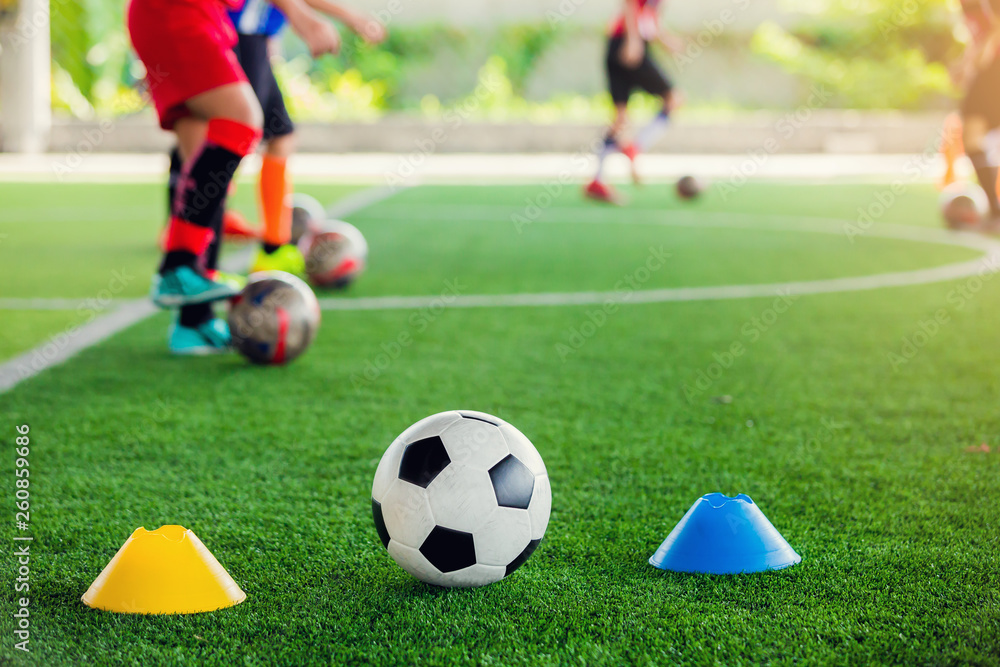 football between marker cones on green artificial turf with blurry soccer team training, blurry kid soccer player jogging between marker cones and control ball with soccer equipment in soccer academy.