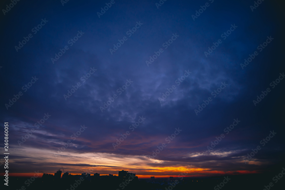 Cityscape with vivid warm dawn. Amazing dramatic blue violet cloudy sky above dark silhouettes of city buildings. Orange sunlight. Atmospheric background of sunrise in overcast weather. Copy space.