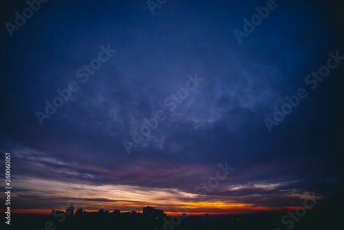 Cityscape with vivid warm dawn. Amazing dramatic blue violet cloudy sky above dark silhouettes of city buildings. Orange sunlight. Atmospheric background of sunrise in overcast weather. Copy space. © Daniil