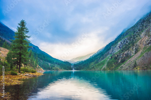 Wonderful mountain lake. Amazing huge mountains with conifer forest. Larch tree on water edge. Morning foggy landscape of majestic nature of highlands. Sunny misty mountainscape. Sunlight through fog.