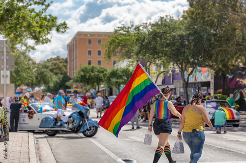 The two girls walk to Pride Fest with a rainbow flag. Walking side by side holding a bag in one hand and the Pride flag poll in the other, they approach the staging area of the parade.