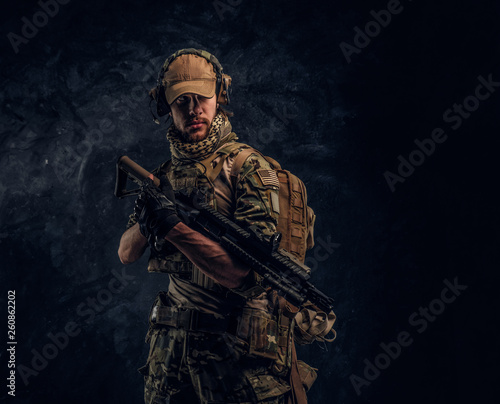 Fully equipped soldier in camouflage uniform holding an assault rifle. Studio photo against a dark wall