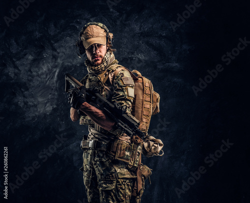 Fully equipped soldier in camouflage uniform holding an assault rifle. Studio photo against a dark wall