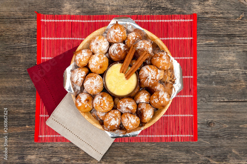 Small homemade buns in a wooden plate. Vanilla sauce in a clay cup and two cinnamon sticks. Red bamboo rug. Kitchen towels of red and natural color. Top view. photo