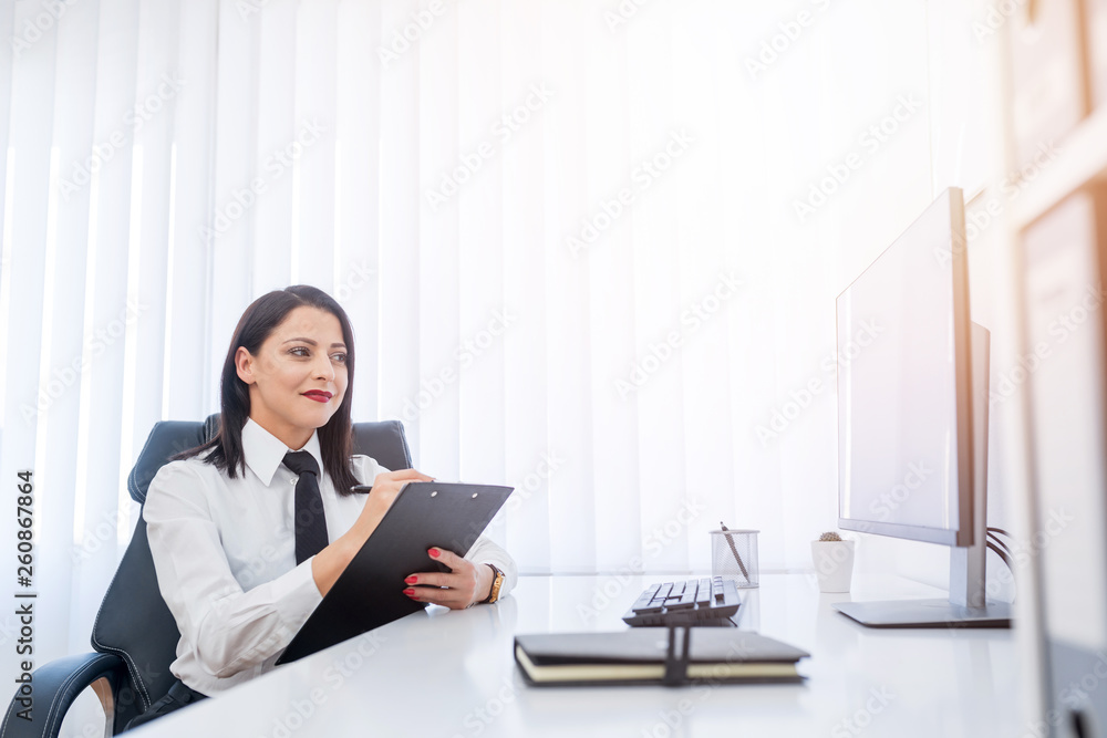 Attractive businesswoman writing information on her clipping board while sitting in her modern office. Attractive businesswoman writing information on her clipping board while sitting in her modern office.