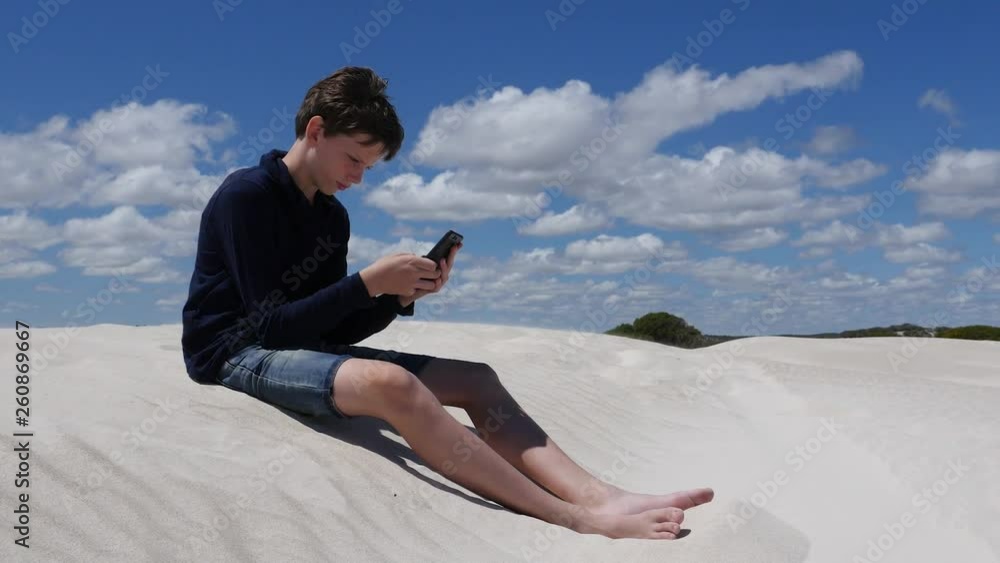 Staying connected in a remote place, boy on dune with smartphone texting