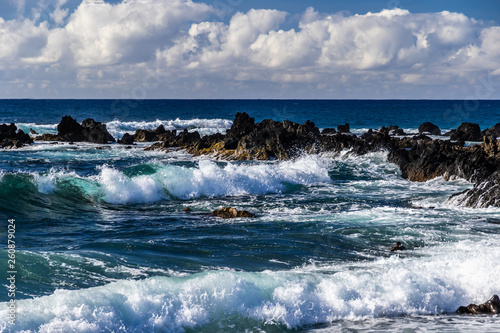 Waves rolling towads shore near South Point, in Hawaii's Big Island. Volcanic rocks in the surf. Blue Pacific Ocean stretches to the horizon; overhead is a sky-filled sky.