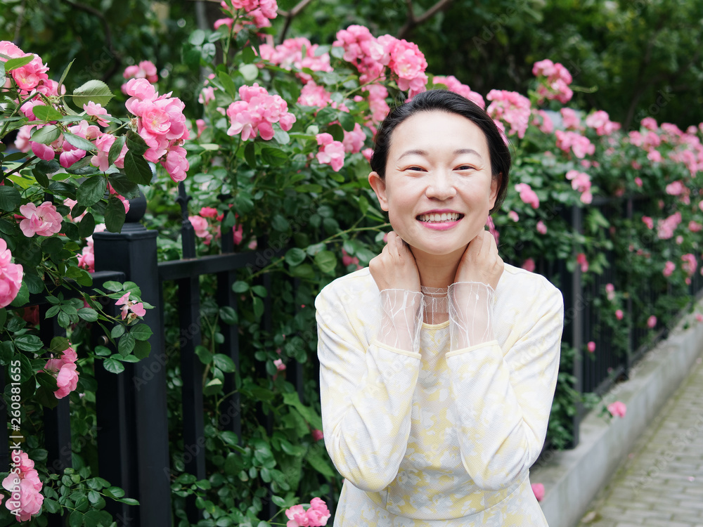 Outdoor portrait of beautiful middle aged Chinese woman in yellow dress smiling among pink rose flowers wall in spring garden, happy mature woman enjoy her leisure time. Outdoor portrait of beautiful middle aged Chinese woman in yellow dress smiling among pink rose flowers wall in spring garden, happy mature woman enjoy her leisure time.