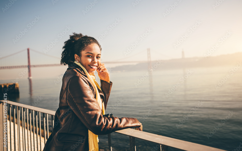 A dazzling cheerful African-American girl is leaning against a fence while standing on the quayside of the Tagus river, enjoying the sunset, with a huge bridge behind and copy space area on the right