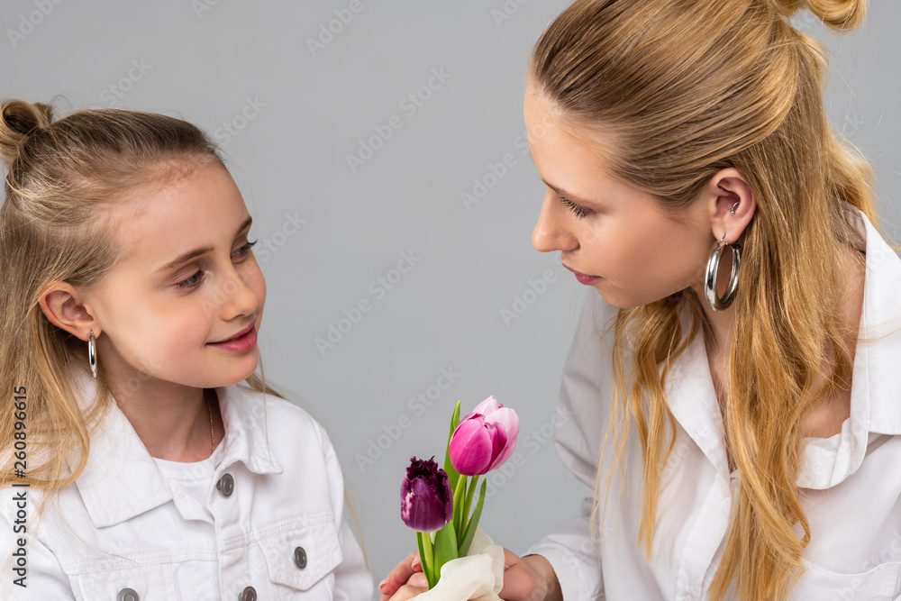 Smiling pretty girl in white jacket gifting flowers to her light-haired sister Smiling pretty girl in white jacket gifting flowers to her light-haired sister