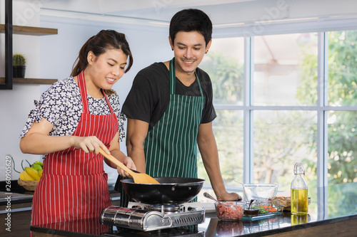 Portrait of happy young and beautiful Asian couple sharing good times preparing lunch together  wife cooking and check omelet in frying pan  in modern kitchen with large glass window green background