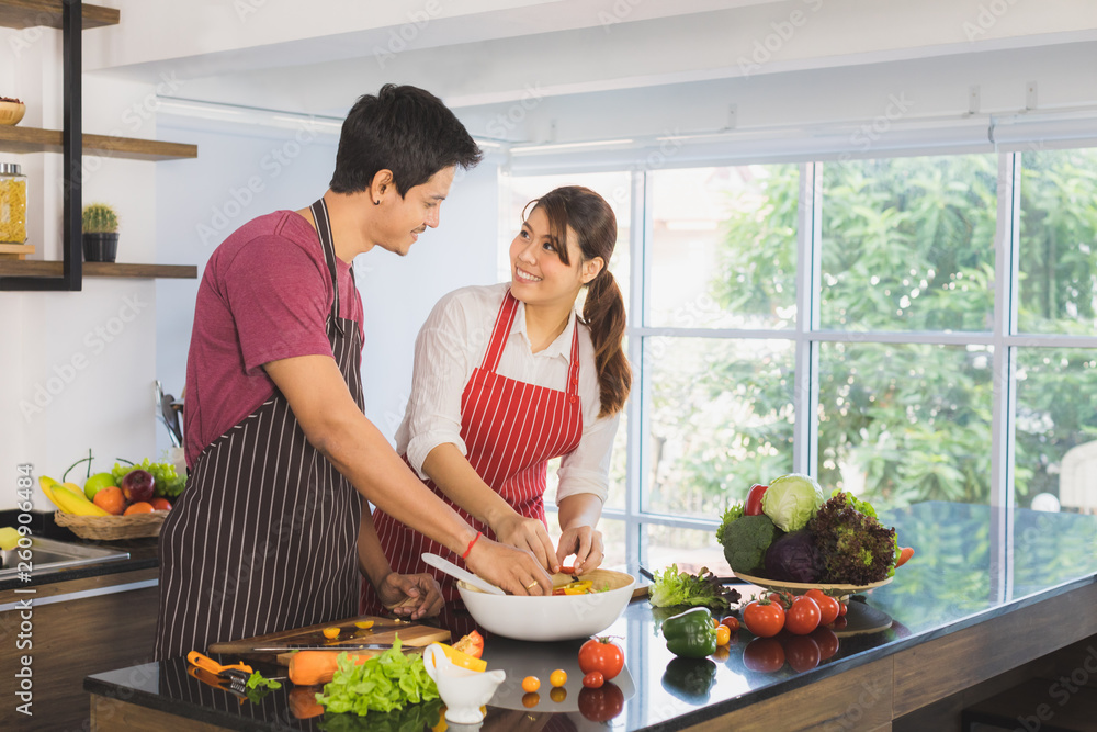 Portrait of happy young Asian couple sharing a good times prepare healthy salad, cutting and slicing vegetable in modern kitchen space with large glass window and blur green garden background Portrait of happy young Asian couple sharing a good times prepare healthy salad, cutting and slicing vegetable in modern kitchen space with large glass window and blur green garden background