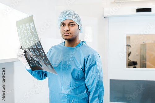 handsome doctor in medical cap and uniform holding x-ray and looking at camera