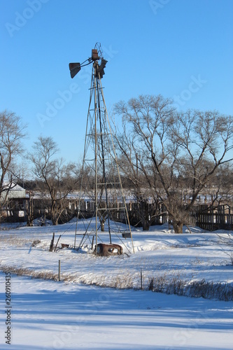 Winter landscape with a old windmill and a old car at its base a couple trees and a old fence.