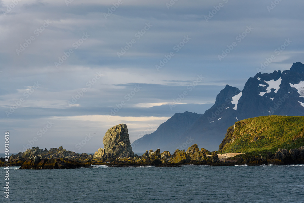 Beautiful landscape of Coopers Bay from the water, rocky spit lit by sunbeam against a moody sky, South Georgia, southern Atlantic Ocean