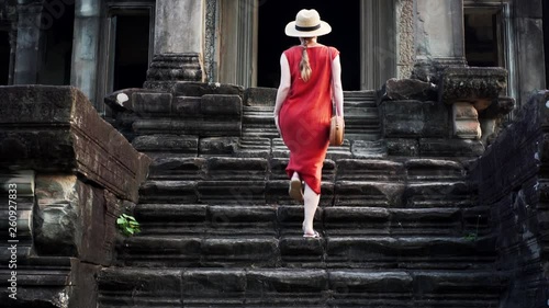 Slow motion: back of woman tourist in straw hat and red dress going upstairs to the Angkor Wat temple entarnce. It was built in 12th century in Cambodia and dedicated to Vishnu photo