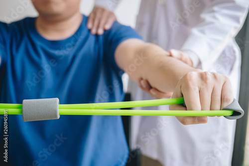 Doctor physiotherapist assisting a male patient while giving exercising treatment on stretching his arm with exercise band in the clinic, Rehabilitation physiotherapy concept