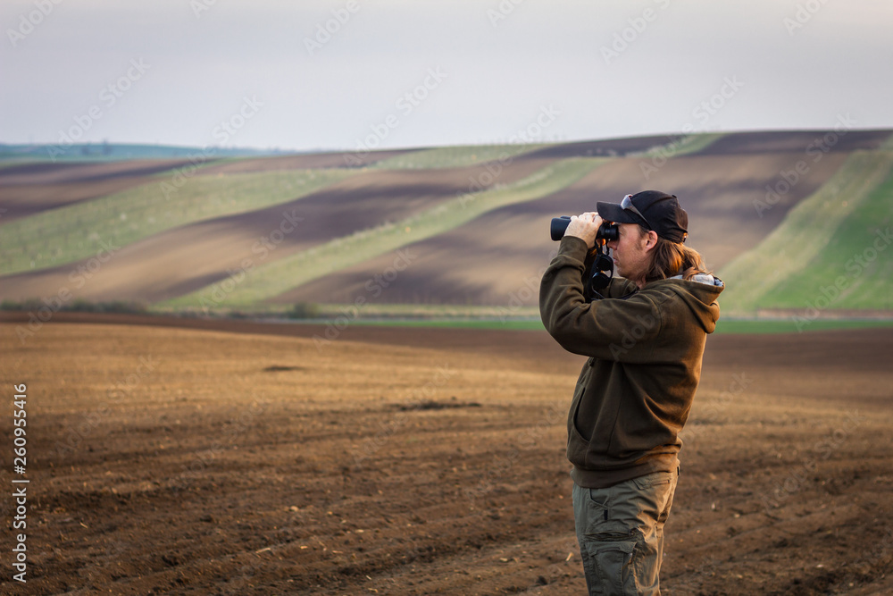 Hiking man looking for animals by binoculars in nature. Birdwatching in rural scene know as Moravian Tuscany, Czech Republic