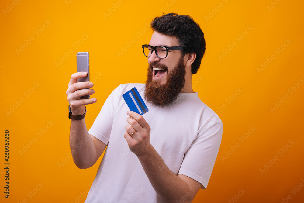 Excited bearded guy using smartphone and credit card for online shop over yellow background Excited bearded guy using smartphone and credit card for online shop over yellow background