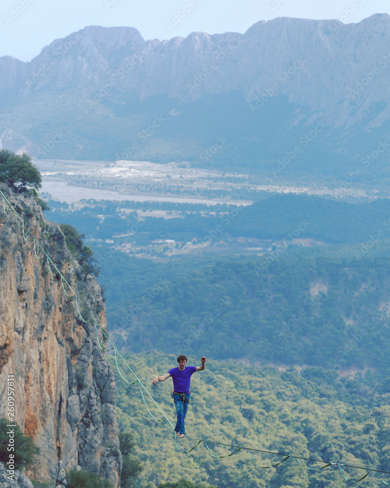 Obraz premium A man is walking along a stretched sling. Highline in the mountains. Man catches balance. Performance of a tightrope walker in nature. Highliner on the background of valley.