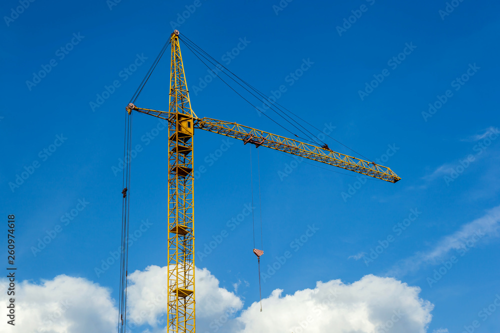 Large yellow construction crane with hook and white clouds with a blue sky on the background. Industrial crane working on a construction site. Large yellow construction crane with hook and white clouds with a blue sky on the background. Industrial crane working on a construction site.