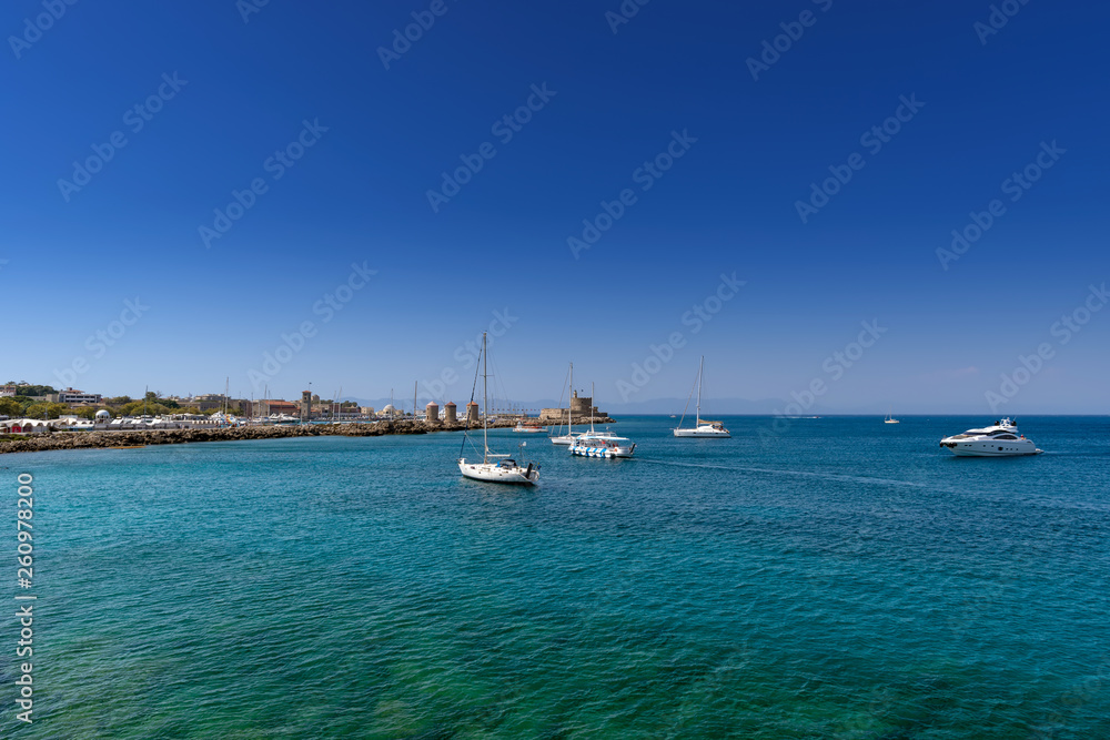 Luxury yachts are anchored in the clear turquoise water near the old Mandraki harbour of the Medieval City of Rhodes in Dodecanese. Famous windmills and Fort of Saint Nicholas in the background.