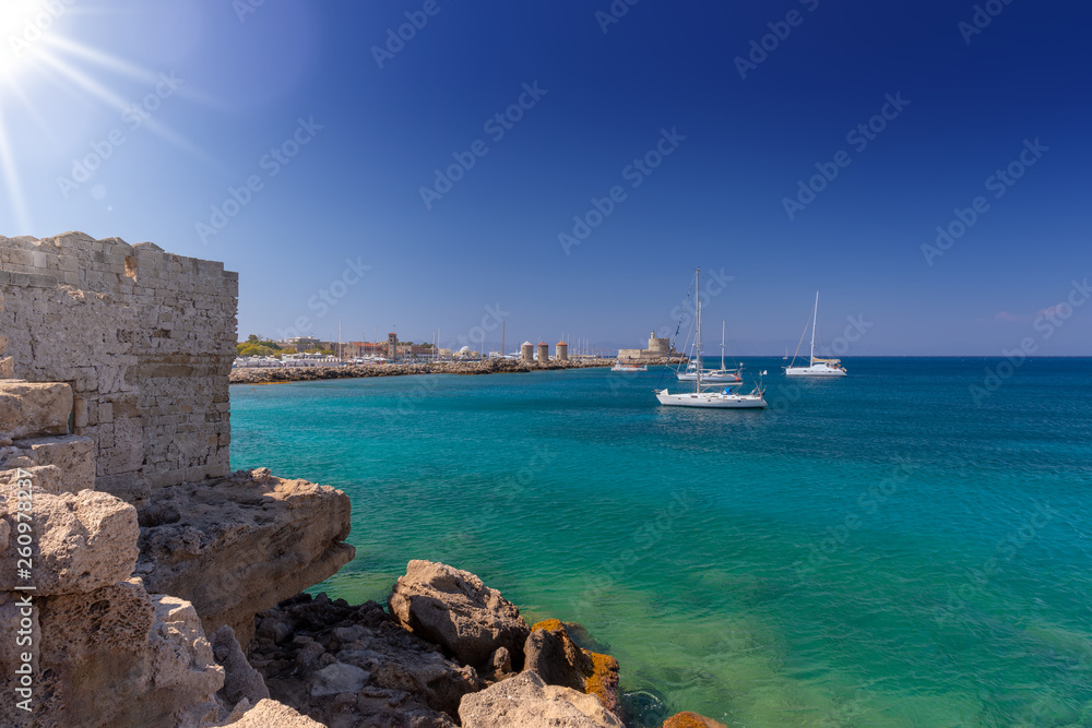 Luxury yachts are anchored in the clear turquoise water near the old Mandraki harbour of the Medieval City of Rhodes in Dodecanese. Famous windmills and Fort of Saint Nicholas in the background.