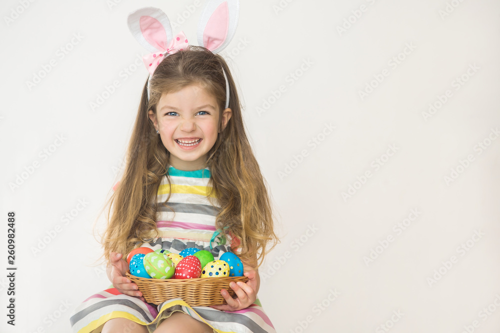 Cute little girl wearing bunny ears holding painted Easter eggs and smiling on a white background.