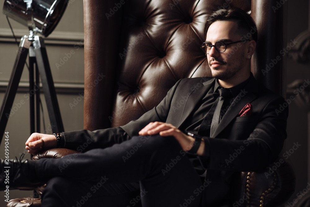 Young handsome man posing for a fashion shoot in a studio. Fashion as a lifestyle. Man wearing a beard. Model sitting on a sofa in tuxedo. Successful fashionable businessman. Business look advertising Young handsome man posing for a fashion shoot in a studio. Fashion as a lifestyle. Man wearing a beard. Model sitting on a sofa in tuxedo. Successful fashionable businessman. Business look advertising