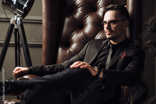 Young handsome man posing for a fashion shoot in a studio. Fashion as a lifestyle. Man wearing a beard. Model sitting on a sofa in tuxedo. Successful fashionable businessman. Business look advertising