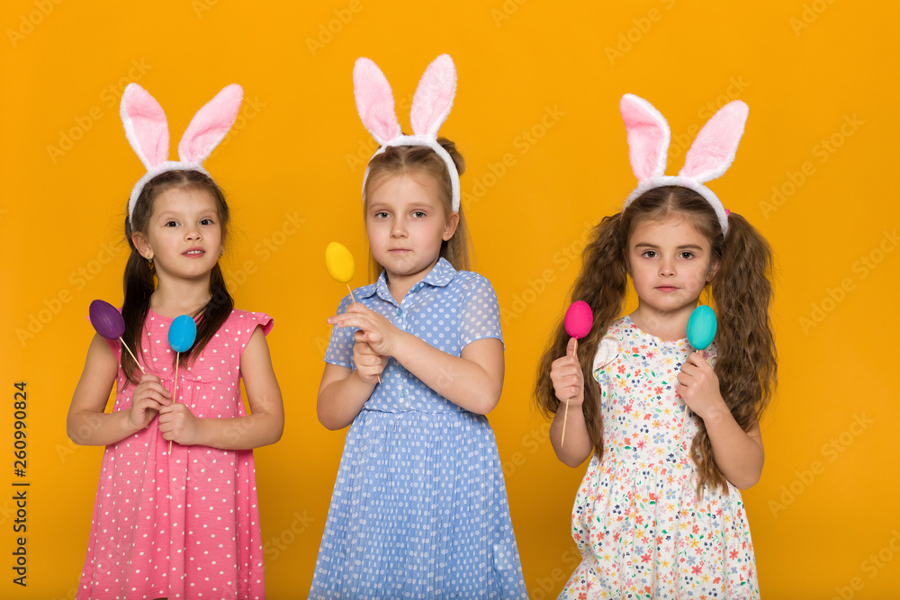 three little girls with Easter bunny ears holding colorful eggs on yellow background. Happy easter. cute children three little girls with Easter bunny ears holding colorful eggs on yellow background. Happy easter. cute children