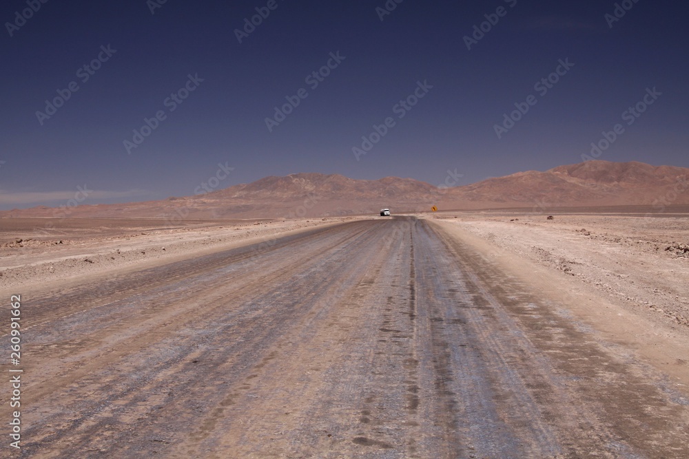 Endless dirt road to infinity of salt flat plateau contrasting with blue cloudless sky. In the horizon isolated truck and yellow traffic sign showing left direction - Atacama desert , Chile