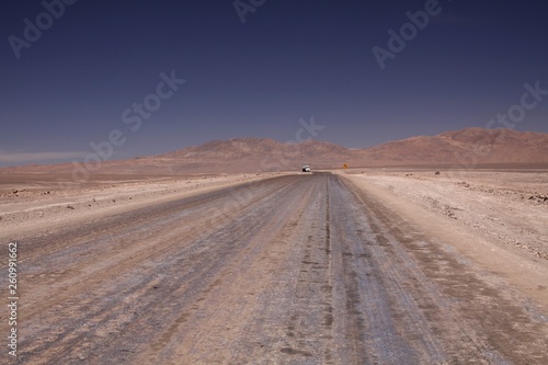 Endless dirt road to infinity of salt flat plateau contrasting with blue cloudless sky. In the horizon isolated truck and yellow traffic sign showing left direction - Atacama desert , Chile