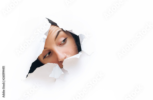 A young girl with a cunning expression. Portrait of a woman looking through the hole in white paper. Distrustful look. Women's curiosity and gossip. A jealous wife. photo