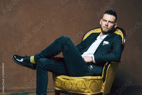 Young handsome man posing for a fashion shoot in a studio. Fashion as a lifestyle. Man wearing a beard. Model sitting on a sofa in tuxedo. Successful fashionable businessman. Business look advertising