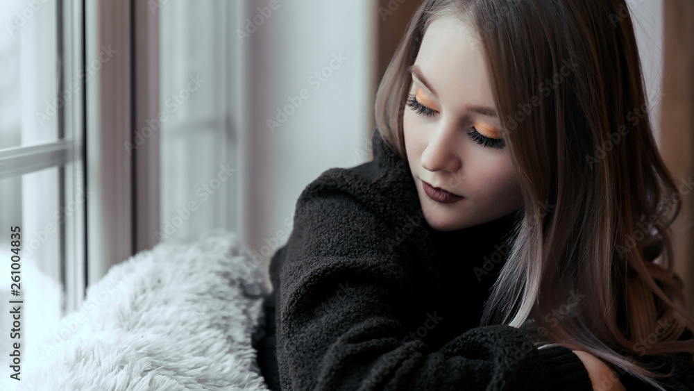 A beautiful young girl is sitting on the windowsill alone with her eyes closed. A woman in sweater sitting by the window A beautiful young girl is sitting on the windowsill alone with her eyes closed. A woman in sweater sitting by the window