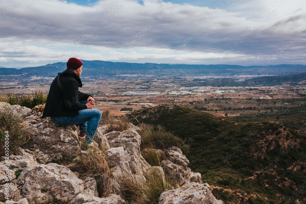 Young male sits on the rock and enjoys beautiful mountain landscape. Small city in background.