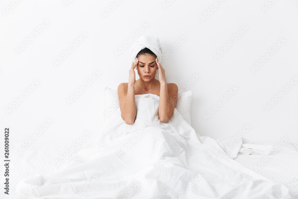 Sad amazing woman with towel on head lies in bed under blanket isolated over white wall background with headache.