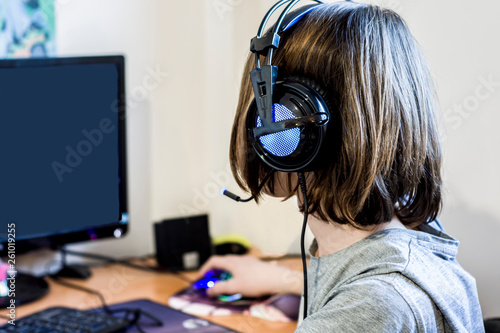 A modern teenager, with a long haircut, plays a computer game in her room at the table against the bright wall in new headphones