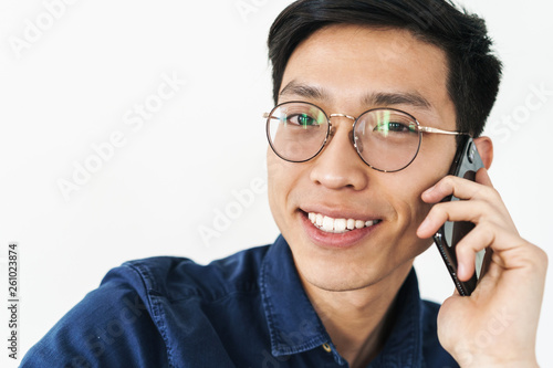 Photo of brunette asian businessman 20s wearing eyeglasses sitting in chair and talking on smartphone while working in office