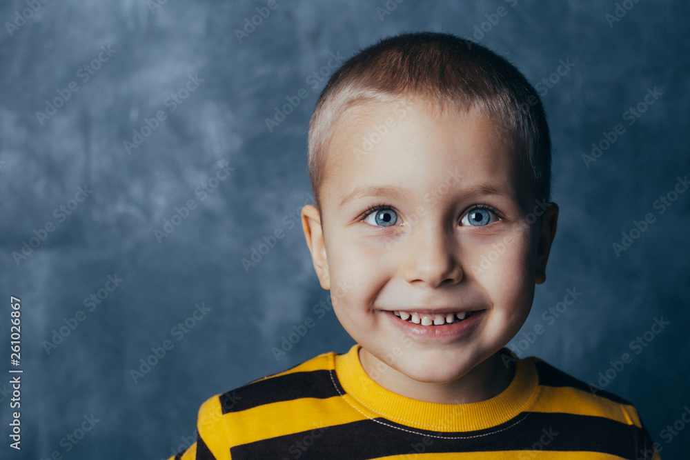 A little boy poses in front of a gray-blue concrete wall. Portrait of a smiling child dressed in a black and yellow striped sweater and brown pants. A little boy poses in front of a gray-blue concrete wall. Portrait of a smiling child dressed in a black and yellow striped sweater and brown pants.