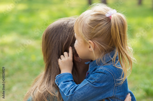 little happy girl hugs her mom and tells her something in the ear in the park