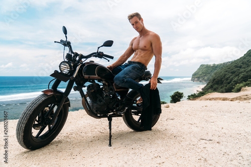 Sexy athletic man with perfect naked body sitting on motorbike, ocean waves and beautiful mountains on background