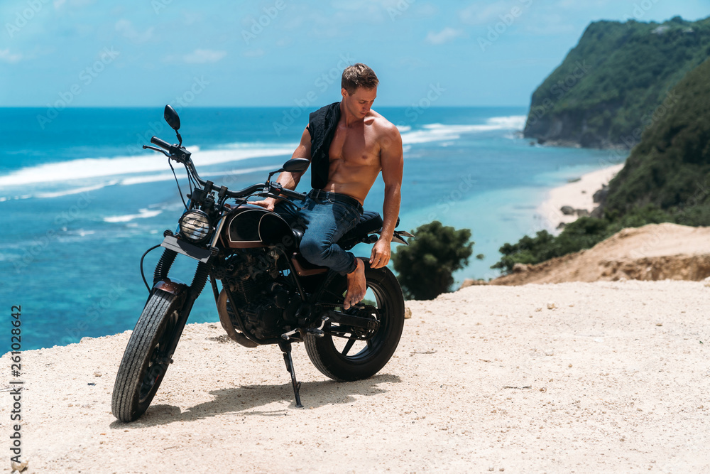 Obraz premium Sexy athletic man with perfect naked body sitting on motorbike, ocean waves and beautiful mountains on background