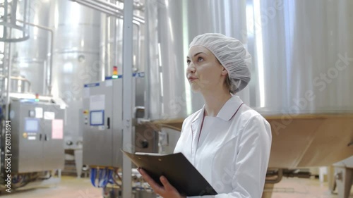 Female Food factory worker monitors production and takes notes on tablet at modern diary factory. Milk processing line
