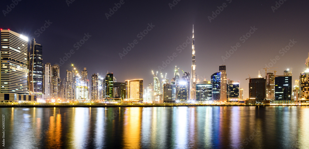 Obraz premium Stunning view of the illuminated Dubai skyline during sunset with the magnificent Burj Khalifa and many other buildings and skyscrapers reflected on a silky smooth water flowing in the foreground.
