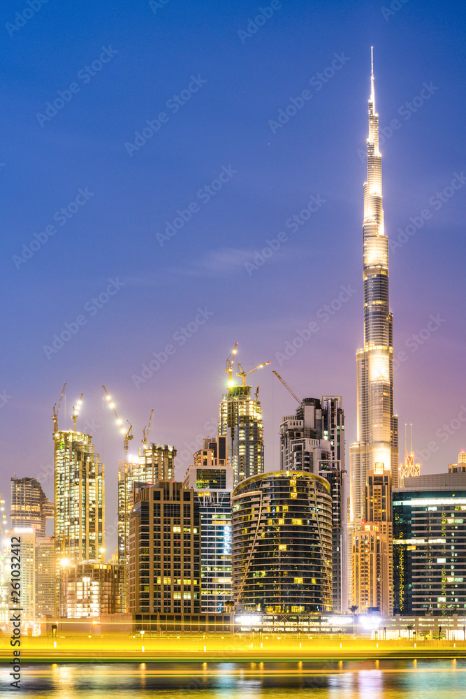 Stunning view of the illuminated Dubai skyline during sunset with the magnificent Burj Khalifa and many other buildings and skyscrapers reflected on a silky smooth water flowing in the foreground.